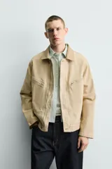 Cazadora relaxed fit de Zara confeccionada en denim de algodón color beige, con cuello de solapa en piel combinada, bolsillos con cremallera en el pecho y cierre frontal oculto.