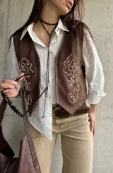 Chaleco de gamuza color marrón chocolate, con diseño sin mangas y escote en V. Cuenta con bordados en hilo claro y pequeños detalles de tachas en la parte frontal.