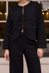 Saco de punto negro con diseño abierto al frente, cierre con lazos de tela y apliques brillantes distribuidos en líneas horizontales. Cuenta con terminaciones onduladas en cuello, puños y ruedo.