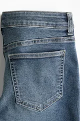 Jeans de denim azul medio con ligero efecto desgastado, tiro bajo y corte acampanado (bootcut) desde la rodilla. Presenta cierre y botón frontal, bolsillos delanteros decorativos y bolsillos abiertos traseros.
