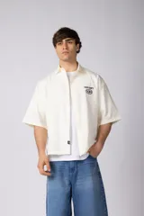 Camisa de manga corta color marfil, confeccionada en 100% algodón de tacto suave, con calce boxy, cuello camisero, cierre frontal de botones y diseño bordado en el pecho.