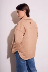 Sobrecamisa negra de estilo shacket, confeccionada en tejido de gamuza sintética. Presenta cuello camisero, cierre frontal de botones y dos bolsillos de parche con solapa en el pecho. El calce es oversize y las mangas son largas.