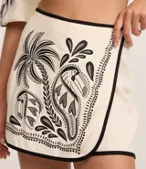 Short pollera de viscolino color crudo con estampa tropical en negro que presenta palmeras y tucanes. Cuenta con cintura fija, cierre posterior con zíper y detalle de vivo contrastante en los bordes.