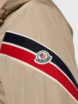 Rompevientos Moncler modelo Claut, color beige, con capucha, cierre frontal y detalle de franjas tricolores en la manga.