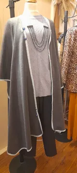 Capa larga gris con vivo blanco en los bordes y botones decorativos. Se cierra con broche en el cuello.