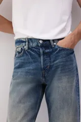 Jeans de cinco bolsillos en denim de algodón rígido, color azul medio con efecto desgastado y lavado. Modelo de cintura clásica con cierre de botón y corte relajado de cadera a bajo con pierna recta más amplia.