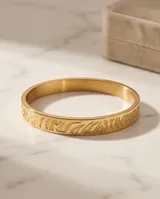Esclava rígida de acero con baño de oro, con diseño grabado en relieve. Se presenta junto a una pulsera de cadena fina de eslabones.