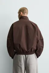 Cazadora técnica de corte boxy fit en color marrón, con cuello subido, cierre frontal de cremallera, bolsillos laterales y uno frontal con cierre, y acabados elásticos en puños y ruedo.