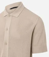 Camisa de punto de manga corta en color beige, con cuello inglés y cierre frontal de botones. Presenta una textura tejida y un corte regular que ofrece un ajuste cómodo y clásico.