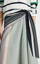 Pollera midi de algodón con silueta en línea A, cintura alta con cruce frontal y lazo lateral. Presenta un estampado de rayas en diferentes anchos y direcciones, con dobladillo recto y pespuntes.
