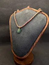 Collar de diseño artesanal compuesto por cordones trenzados multicolor en tonos verdes, rosados y rojizos. Presenta un dije de piedra ovalada con una cruz dorada aplicada en el centro.