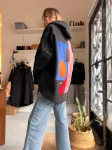 Campera de textura tipo corderito en color negro, con diseño de collage geométrico en la espalda que incluye parches en azul, naranja, marrón y franjas rojas y blancas. Posee capucha y mangas largas.