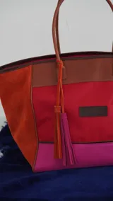 Bolso de fin de semana tipo tote, confeccionado en una combinación de lona y gamuza en tonos rojo, fucsia y marrón. Cuenta con doble asa de hombro, cierre superior y detalle de borlas colgantes en el lateral.