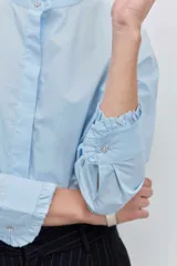 Camisa de algodón color celeste, con cuello mao y puños con detalle de volados. Presenta cierre frontal oculto con botones y un diseño de corte clásico.