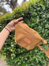 Riñonera de gamuza color tostado con cierre frontal y etiqueta de cuero con logo de la marca Antonia Wool Wear. Cuenta con correa ajustable y herrajes metálicos.