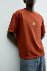 Camiseta de manga corta y cuello redondo, color terracota, con un pequeño bordado de una flor amarilla y la inscripción "Abstract Harmony" en el pecho.