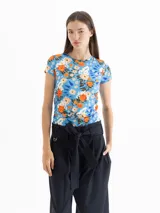 Top de punto color azul con estampado floral en tonos naranja, blanco y verde. Tiene escote redondo, mangas cortas y frunces laterales que estilizan la figura.