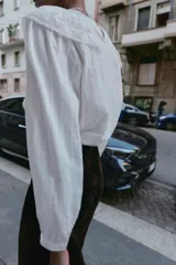 Camisa blanca de cuello redondo y manga larga, con detalle de volantes frontales y entredoses de encaje. Presenta un cierre en la espalda con abertura y botón.