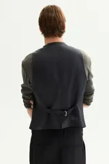 Chaleco de traje negro con cierre frontal de cinco botones y bolsillos ribeteados. Presenta un corte entallado, forro interior y espalda ajustable con trabilla.