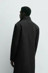 Abrigo negro de corte recto con cuello subido y doble cuello desmontable mediante cremallera interior. Cuenta con manga larga, bolsillos de vivo en la cadera, bolsillo interior, abertura central en la espalda y cierre frontal de cremallera oculta por solapa con botones a presión.