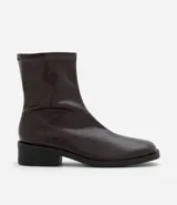 Bota tipo sock boot de caña media, confeccionada en poliuretano color marrón oscuro con acabado brillante. Presenta puntera redondeada, cierre lateral y taco bloque bajo de color negro.