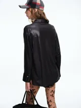 Camisa de corte oversize confeccionada en tejido jersey con acabado brillante. Presenta cuello en punta, cierre frontal con botones, mangas largas con puños anchos y tajos laterales en el ruedo.