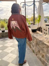 Campera bomber de jean color terracota, con corte holgado. Presenta apliques de cuero cortados y bordados a mano en la espalda, delantera y cuello, formando un diseño de montañas en tonos beige, rosa y verde militar.