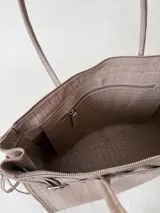 Bolso grande de cuero marrón con textura tipo cocodrilo, estructura rígida y asas para llevar al hombro. Tiene una tira con tachas y hebillas en cada extremo, también lleva lazos en los costados.