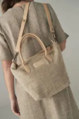 Cartera tipo bolso de mano confeccionada en lana fina sin teñir, con detalles en cuero marrón oscuro en las asas y correa ajustable. Incluye cierre superior y etiqueta de cuero con logo.