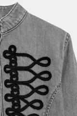 Chaqueta de denim gris con cuello subido y manga larga. Presenta un diseño entallado con bajo en evasé y un llamativo bordado frontal de alamares en color negro. Cierre frontal con corchetes ocultos.