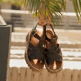 Sandalias de cuero vacuno en color negro, con diseño de tiras cruzadas, suela track de goma y tira ajustable con hebilla en el tobillo.