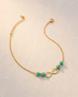 Tobillera de cadena fina de acero con baño de oro, con dije central de símbolo de infinito y cuentas de piedra turquesa.
