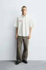 Camisa de algodón blanca con finas rayas verticales negras, corte relajado (relaxed fit) y manga corta. Presenta un bordado en hilo rojo en el lado izquierdo del pecho.