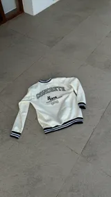 Buzo color crema con cuello, puños y cintura acanalados con rayas negras. Estampado en la espalda con la palabra "CONCRETE" en mayúsculas, el número "1978", un conejo y la frase "SOUND & VISUAL PROGRAM".