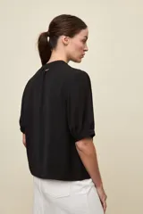 Blusa de manga corta color negro, con calce relajado, escote redondo con abertura y lazo para anudar. Presenta detalles de tablas horizontales en la parte superior que aportan textura al diseño.