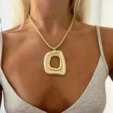 Collar con cordón trenzado color beige y dije grande de metal dorado con forma orgánica irregular y agujero central.