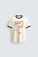 Camiseta de corte regular fit, color crema, con cuello redondo y mangas cortas. Presenta un estampado frontal con un personaje de dibujos animados jugando al baloncesto y texto "Hudson Valley Company Champion '87 NYC, USA". Los acabados del cuello y las mangas son de vivo negro a contraste.