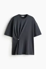 Remera de algodón de corte holgado con hombros caídos y cuello redondo. Presenta un detalle metálico decorativo en un lateral que genera un efecto drapeado.