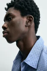 Camisa de hombre de manga larga, color azul, con cuello solapa y bolsillo plastrón en el pecho. Presenta un patrón de cuadros pequeños en tonos de azul y blanco.