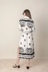 Vestido camisero midi de lino y viscosa color crudo con estampado de palmeras en negro. Tiene cuello camisero, mangas largas con puños y cinturón de la misma tela.