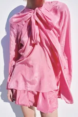 Blusa fluida de color rosado con cuello redondo ajustable mediante lazadas del mismo tejido, manga larga con puño y bajo asimétrico con aberturas laterales.
