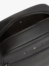 Bandolera reporter Tommy Hilfiger de color negro, confeccionada en poliuretano con acabado liso y panel frontal inferior con detalle tejido. Cuenta con un compartimento principal con cierre de cremallera, bolsillo exterior frontal también con cremallera, bolsillo interior abierto, correa ajustable y logo metálico de la marca en la parte delantera.