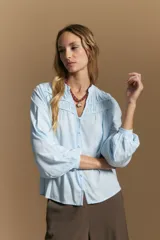 Camisa de manga larga color celeste, con cuello mao y escote en V. Presenta frunces en el canesú, cierre frontal con botones y mangas amplias con puños abotonados.