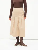 Falda midi de color beige, con diseño de tablas frontales y cierre lateral con botones decorativos. Presenta un corte estructurado y tiro alto.