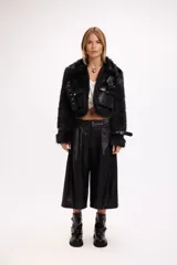 Campera corta de pelo sintético negro con detalles en cuero sintético, incluyendo bolsillos frontales con solapa, cinturón con hebilla metálica en el pecho y correas ajustables en los puños.