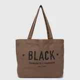 Bolso tipo tote marrón de lona con doble asa de hombro y estampado frontal con la palabra "BLACK" y texto descriptivo.