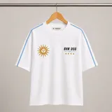 Remera blanca de corte holgado con detalles de líneas celestes en los hombros y mangas. Presenta bordados frontales: un sol de estilo patrio a la izquierda y el texto BHN DSG junto a cuatro estrellas doradas a la derecha.