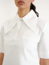 Camisa blanca de manga corta con cuello camisero grande y marcado, cierre frontal oculto y bolsillo ojal en el pecho.