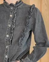 Camisa de denim celeste con cuello alto con volados, botonadura frontal y detalles de volados verticales a lo largo del frente. Cuenta con mangas largas abullonadas y puños con volados.