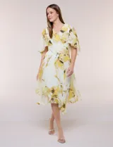 Vestido midi de gasa liviana con estampado floral en tonos amarillo y blanco, escote en V cruzado, mangas cortas amplias tipo capa, cintura entallada y falda con vuelo y ruedo irregular.